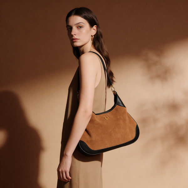 A woman in a beige sleeveless dress carries a large brown suede hobo shoulder bag with black leather trim and a gold-tone logo clasp, set against a warm beige background with artistic shadows.